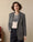Una donna indossa un blazer monopetto in lana grigia motivo spigato, abbinato a un maglione bianco con pattern geometrico blu e pantaloni scuri.