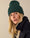 Una donna indossa un cappotto in lana blu scuro abbinato a un berretto a coste verde, su sfondo neutro.